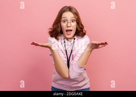 Porträt von geschockt junge Brünette Teenager Mädchen in Brillen werfen Hände nach oben und zuckende Schultern, suchen mit großen Augen und offenen Mund. Innen Stockfoto