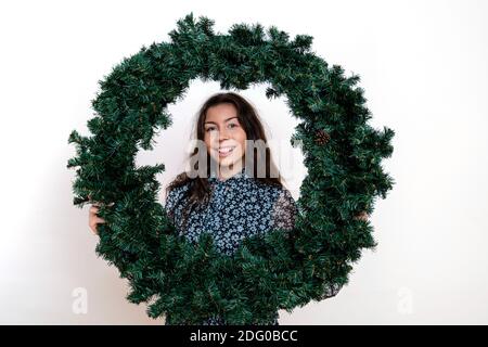 Gut aussehende Brünette Frau schaut durch Weihnachtskranz, isoliert auf weißem Hintergrund Stockfoto