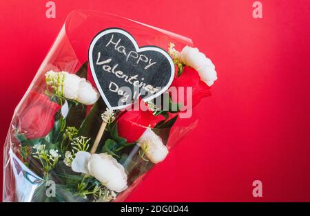 Blumenstrauß aus roten und weißen Rosen, mit einem Tafelherz mit der in Kreide geschriebenen Botschaft 'Happy Valentine's Day' vor rotem Hintergrund. Stockfoto