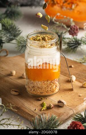 Kürbisparfait mit Pistazien in Glas Mason Jar Becher auf einem Holzbrett vor dem Hintergrund der Neujahrsdekorationen, Pistazien fallen von oben. Stockfoto