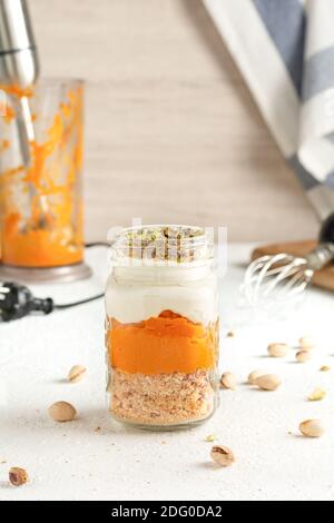 Kürbis Parfait mit Pistazien in Glas Mason Becher. Im Hintergrund ein Mixer mit übrig gebliebenes Kürbispüree, ein Blätterteig und ein blau gestreiftes T Stockfoto