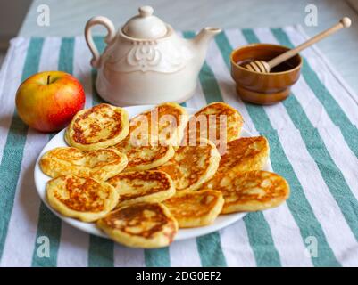 Leckere hausgemachte Pfannkuchen auf dem Teller in der Nähe eines Apfels, Teekanne und Schüssel mit Honig gelegt. Selektiver Fokus auf Pfannkuchen Stockfoto