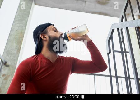 Seitenansicht des aktiven bärtigen Sportlers, der Wasser aus der Glasflasche trinkt, während er sich ausruhte und an einem hellen Tag auf der Treppe saß. Stockfoto