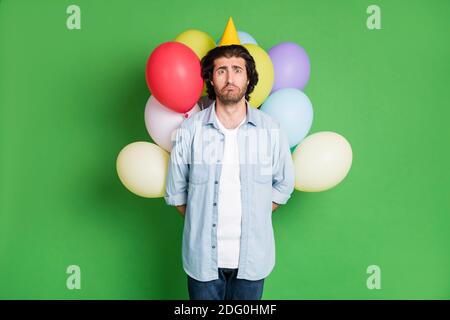 Foto von Mann halten Ballons hinter Rücken unglücklich Blick Kamera Tragen Papier Kegelkappe Denim Hemd Jeans isoliert grüne Farbe Hintergrund Stockfoto