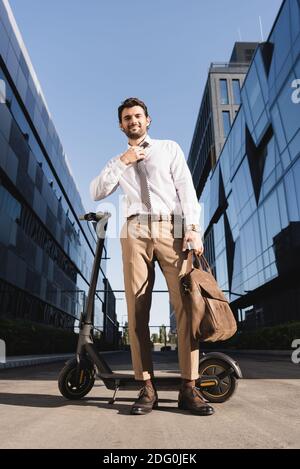 Low-Winkel-Ansicht der bärtigen Geschäftsmann in formellen tragen Anpassung Krawatte, während die Ledertasche hält und in der Nähe von Elektroroller steht Stockfoto
