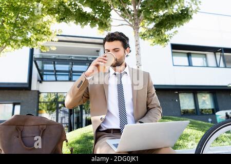 Geschäftsmann in schnurlosen Ohrhörern und Anzug trinken Kaffee zu gehen Und mit Laptop auf der Bank sitzen Stockfoto