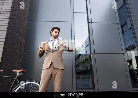 Bärtiger Geschäftsmann hält Kaffee zu gehen und Smartphone während der Suche Bei der Uhr in der Nähe des Fahrrads Stockfoto