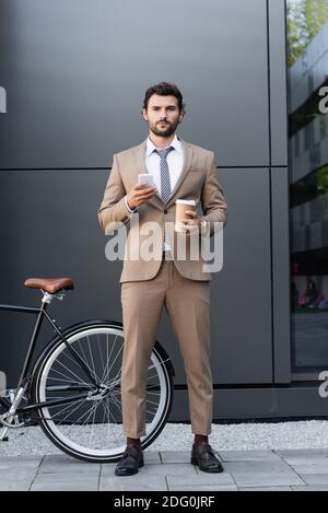 Volle Länge der bärtigen Geschäftsmann hält Kaffee zu gehen und Smartphone in der Nähe des Fahrrads Stockfoto