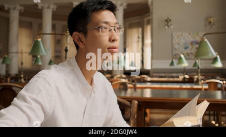 Portrait eines asiatischen Studenten mit Lehrbuch traumhaft wegschauen in der Bibliothek. Junger Mann, der an der Universität studiert Stockfoto