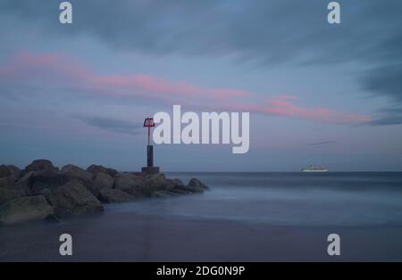 Ein Kreuzfahrtschiff vor der Küste aufgrund Covid-19 liegt am Horizont eines Meeres ruhig durch eine lange Exposition unter einem Band von rosa Wolken bei Sonnenuntergang gemacht. Stockfoto