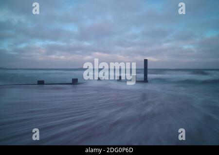 Wellen hinterlassen Spuren auf dem Sand, während sie vom Ufer wegeilen. Unterdessen stürzt das Meer auf das verwitterte Holz der trotzigen Seeverteidigungsgroyne Stockfoto