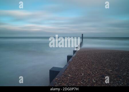 Eine hölzerne Seeverteidigungsgroyne-Markierung führt ins Meer hinaus und teilt die Szene in halb freiliegende Muscheln, halb Meerwasser, das durch eine lange Belichtung geglättet wird. Stockfoto