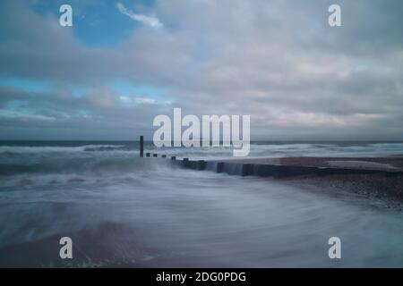 Ein Meeresschutzgroyne schneidet durch die brechenden Wellen eines wütenden Meeres, Wasser rauscht über den Wald und über den Sand, während graue Wolken den Himmel füllen. Stockfoto