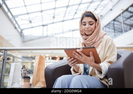 Porträt einer modernen Frau aus dem Nahen Osten, die Kopftuch trägt und ein digitales Tablet benutzt, während sie im Einkaufszentrum sitzt und Platz für Kopien verwendet Stockfoto