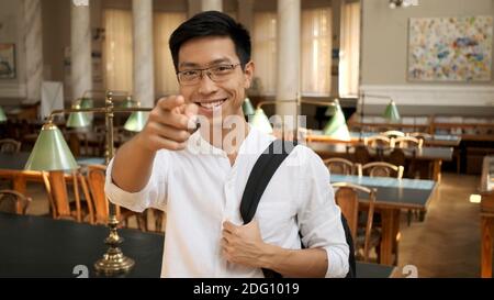 Portrait von fröhlichen asiatischen Schüler selbstbewusst Zeigefinger in der Kamera zeigen. Junger Mann mit Rucksack in der Bibliothek stehen Stockfoto