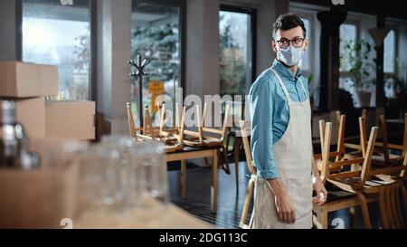 Frustrierter Kellner mit Schürze im geschlossenen Cafe, kleine Geschäfte wurden wegen des Coronavirus geschlossen. Stockfoto