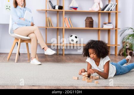 Fokussiertes afroamerikanisches Mädchen, das mit hölzernen Blöcken spielt, während es liegt Auf dem Boden mit verschwommenem Büro im Hintergrund Stockfoto