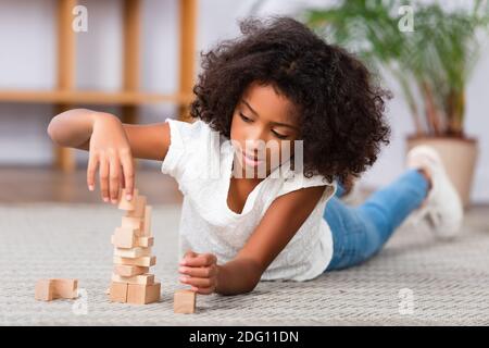 Konzentriertes afroamerikanisches Mädchen, das beim Liegen mit Holzblöcken spielt Auf dem Boden mit verschwommenem Büro im Hintergrund Stockfoto