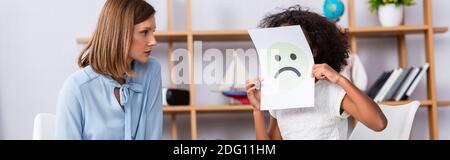 afroamerikanisches Mädchen mit Autismus, das Gesicht mit unglücklicher Expression auf Papier bedeckt, nahe Psychologe auf verschwommenem Hintergrund, Banner Stockfoto