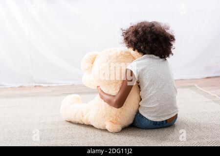 Rückansicht des einsamen afroamerikanischen Mädchens mit Autismus Umarmung teddybär beim Sitzen auf dem Boden in der Nähe des Vorhangs Stockfoto