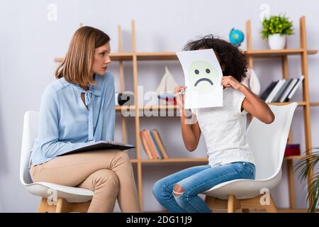 Psychologe Blick auf afroamerikanische Mädchen Abdeckung Gesicht mit unglücklich Ausdruck auf Papier mit verschwommenem Büro im Hintergrund Stockfoto