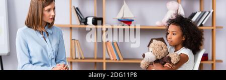 Weibliche Psychologin Blick auf verärgert afroamerikanische Mädchen umarmt Teddybär während der Beratung im Büro, Banner Stockfoto