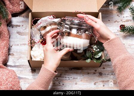Weibliche Hände halten Weihnachts-Geschenkbox mit Cookie-Mix und Schokoladengetränk in Glas auf dunklem Holztisch. Schichten von Mehl, Kakaopulver, Zucker. ICH Stockfoto