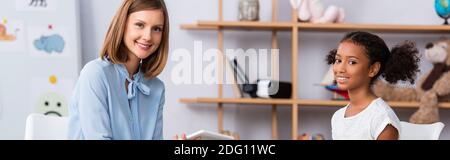 Lächelnde Psychologin und afroamerikanische Mädchen, die während der Konsultation mit verschwommenem Büro auf dem Hintergrund, Banner auf die Kamera schauen Stockfoto