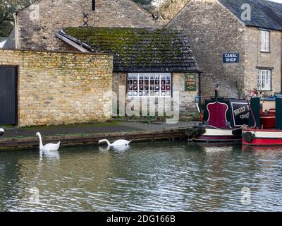 Winterszene auf dem Grand Union Kanal in Stoke Bruerne, Northamptonshire, UK; festgemacht Schmalboot, Kanal-Shop mit Weihnachtsfenster und zwei Schwäne. Stockfoto