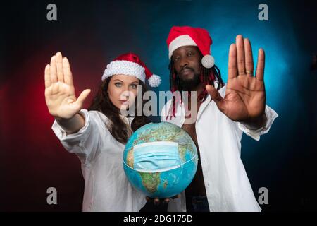 Ein schwarzer Mann und eine europäische Frau in Weihnachtsspaten, die einen Glossen auf seiner Schutzmaske halten. Hände nach oben, Geste gegen. Konzept, kein Coronavirus. Foto im Studio, schwarzer Hintergrund. Hochwertige Fotos Stockfoto