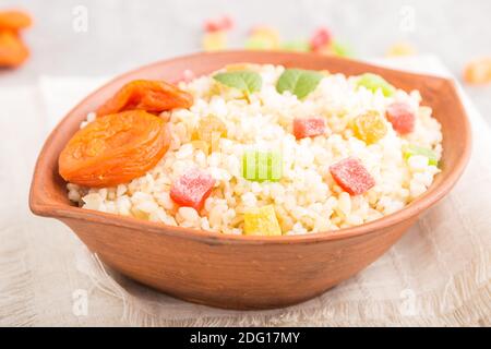 Bulgur-Brei mit getrockneten Aprikosen und kandierten Früchten in Tonschüssel auf grauem Betonboden und Leinentextilien. Seitenansicht, Nahaufnahme, selektive Ansicht Stockfoto