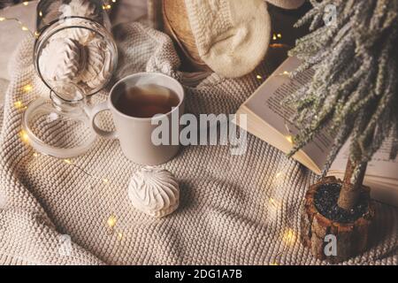 Warmes gemütliches Zuhause zu weihnachten - Tasse heißen Tee, Zephyren und Buch mit Lichtern auf Strickdecke Stockfoto