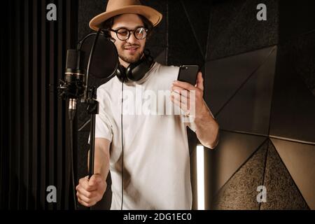 Junge attraktive männliche Musiker Lesung Song Text auf Smartphone Vorbereitung Singen im Tonstudio Stockfoto