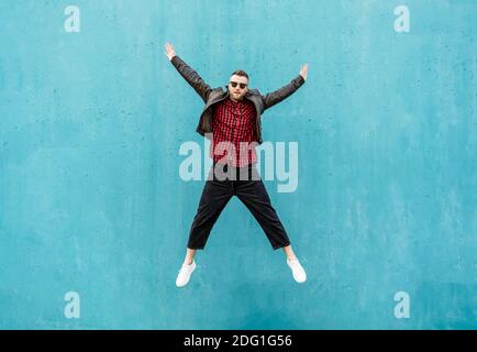 Cool Mann in lässigen Indie Kleidung springen im Freien mit offen Arme vor einer blauen Wand Hintergrund - trendy Kerl mit Spaß beim Tanzen und Feiern im Freien Stockfoto