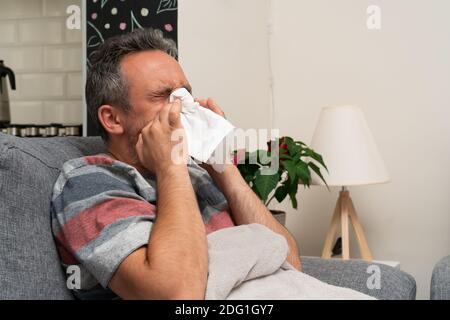 Kranker Erwachsener Mann sitzt auf der Couch mit Grippe sars kalt Influenza covid Symptome Blasen stickige Nase mit Serviette als Pandemie Sinusitis Konzept Stockfoto