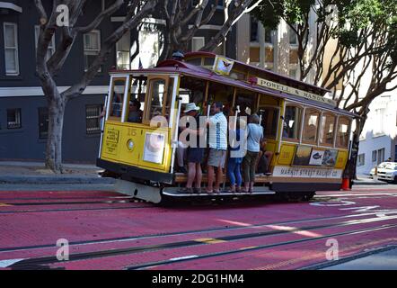 Eine gelbe und rote Cable Car-Linie der Powell & Mason Linie in der Innenstadt von San Fransisco, Kalifornien. Die Seilbahn ist voll von Menschen, die an den Seiten stehen. Stockfoto