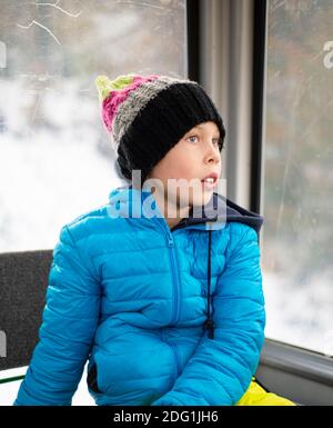 Ein Junge in einer blauen Jacke und einem gestrickten Hut sitzt in einer Kabine am Winterfenster und träumt. Seine blauen Augen blicken in die Ferne. Nachdenklicher Blick. Stockfoto