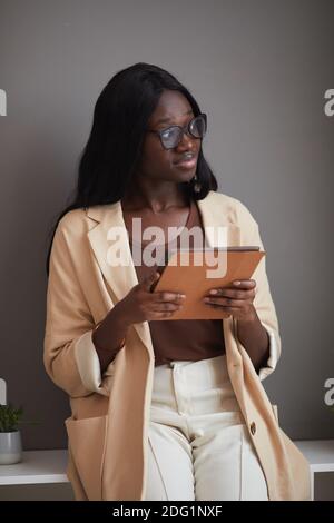 Vertikale Porträt der eleganten afroamerikanischen Geschäftsfrau halten digitale Tablette während Lehnt sich bei der Arbeit an die graue Wand Stockfoto