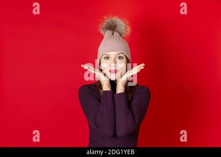 Nahaufnahme einer lächelnden jungen Frau in einem gestrickten Winterhut und Händen in der Nähe ihres Gesichts, isoliert auf einem roten Hintergrund Stockfoto