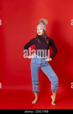 Eine in voller Länge lächelnde junge Frau steht in einem gestrickten Winterhut und Jeans, isoliert auf einem roten Hintergrund Stockfoto