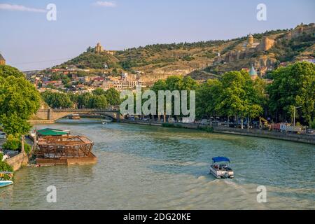 Tiflis, Georgien - 19. Juli 2019: Sommer Stadtbild bei Sonnenuntergang - Panoramablick mit Kura Fluss, bunte traditionelle Häuser, alte Brücke, touristische Boot Stockfoto