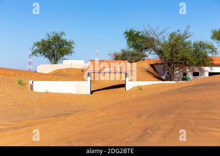 Weißes Eingangstor im Al Madam Geisterdorf, mit Sanddünen und wilden Ghafenbäumen, im Sand begraben. Stockfoto