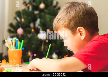 7 Jahre alter Junge, der am Schreibtisch sitzt und stumpf wird. Kind schreibt Notizen in Notizbuch, Nahaufnahme, Seitenansicht. Geschmückter Weihnachtsbaum im Hintergrund Stockfoto