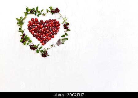 Das Herz ist flach ausgekleidet frische rote Preiselbeeren, gerahmt ein Zweig Kranz aus kastanienbraunen Preiselbeere mit Beeren und grünen Blättern. Auf einer weißen strukturierten Matte Stockfoto