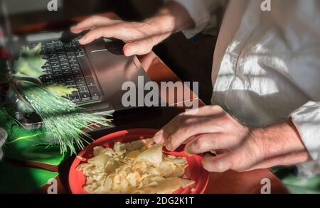 Die Hände eines Mannes, der an seinem Computer arbeitet. Er ist zu Hause, umgeben von Dekorationen für die nächsten Feiertage. Er gönnt sich eine Freude. Stockfoto