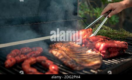 Hand des Mannes mit Metall tong Kochen Schweinefleisch Rippen mit roten Paprika auf BBQ Grill. Kochen Sie ein köstliches gegrilltes Fleisch. Essen gekocht mit Grillen Grill. Koch c Stockfoto