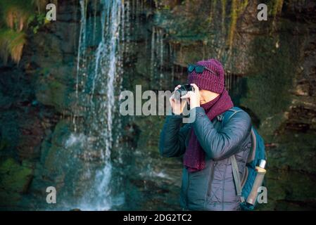 Junger Mann, der während seines Spaziergangs in der Nähe des Wasserfalls mit einer analogen Kamera fotografiert. Lokale Selbstreisen im Herbst, Winter. Hobby-Konzept. Selektiver Fokus. Kopieren Stockfoto