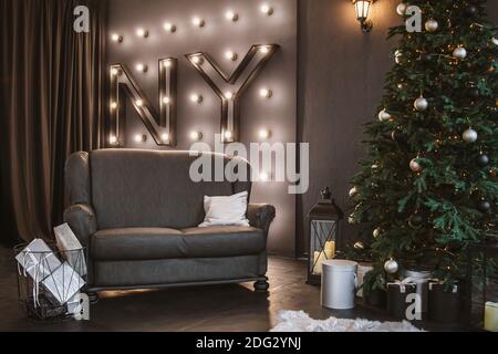 Schöne graue Sofa, Weihnachtsbaum mit Dekorationen, Korb mit Geschenken und Buchstaben an der Wand Neujahr. Gemütliche weihnachtliche Atmosphäre. Stilvolles Zimmer inte Stockfoto