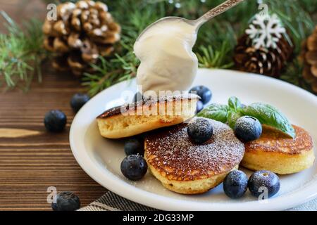 Flauschige heiße Pfannkuchen mit Blaubeeren und Zuckerpulver und dicker Sauerrahm auf dem weißen Teller. Weihnachten Holz Hause Hintergrund aus nächster Nähe. Stockfoto