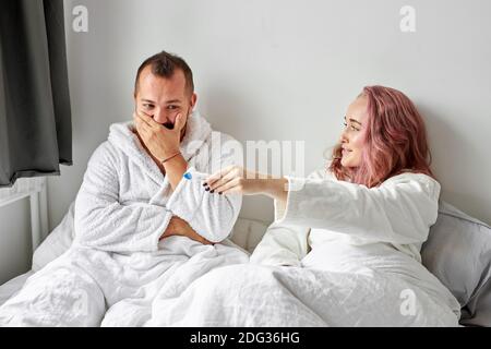 Glückliches kaukasisches Paar, das mit positivem Schwangerschaftstest am Morgen auf dem Bett aufgeregt wird, glückliche Reaktion, ist Mann überrascht Stockfoto